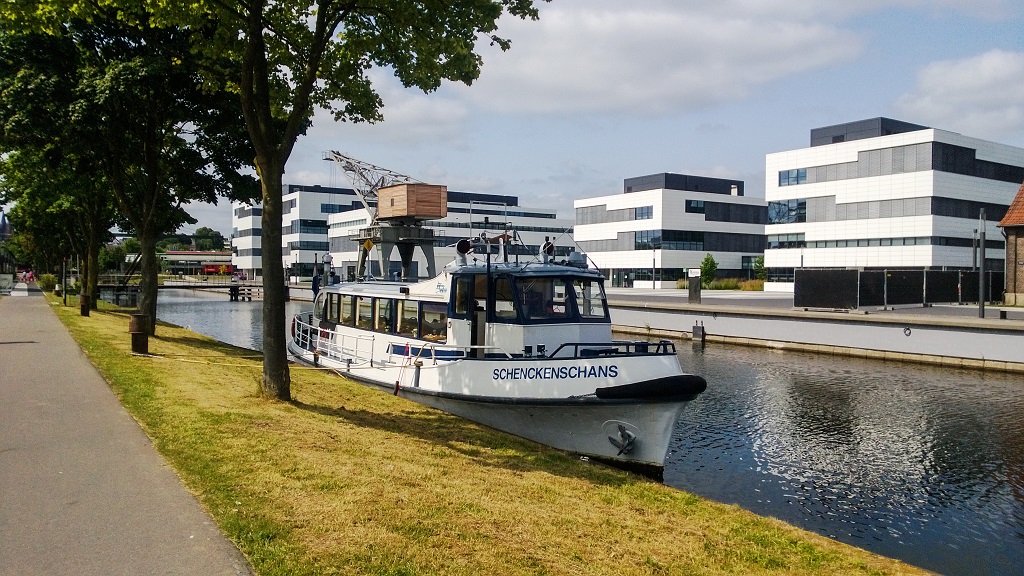 Rondvaartboot Schenckenschans aangemeerd langs het kanaal, met moderne kantoorgebouwen op de achtergrond en bomen langs het wandelpad.