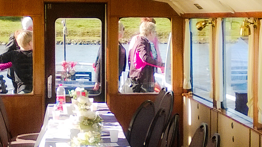 Gedekte tafel met bloemen en servies in de salon van een rondvaartboot, met mensen die buiten op het dek staan en door de ramen zichtbaar zijn.