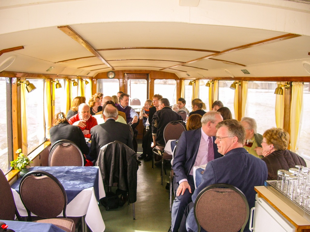 Groep mensen zit samen in de salon van een rondvaartboot tijdens een bijeenkomst, aan tafels met gedekte plaatsen en grote ramen langs de zijwanden.
