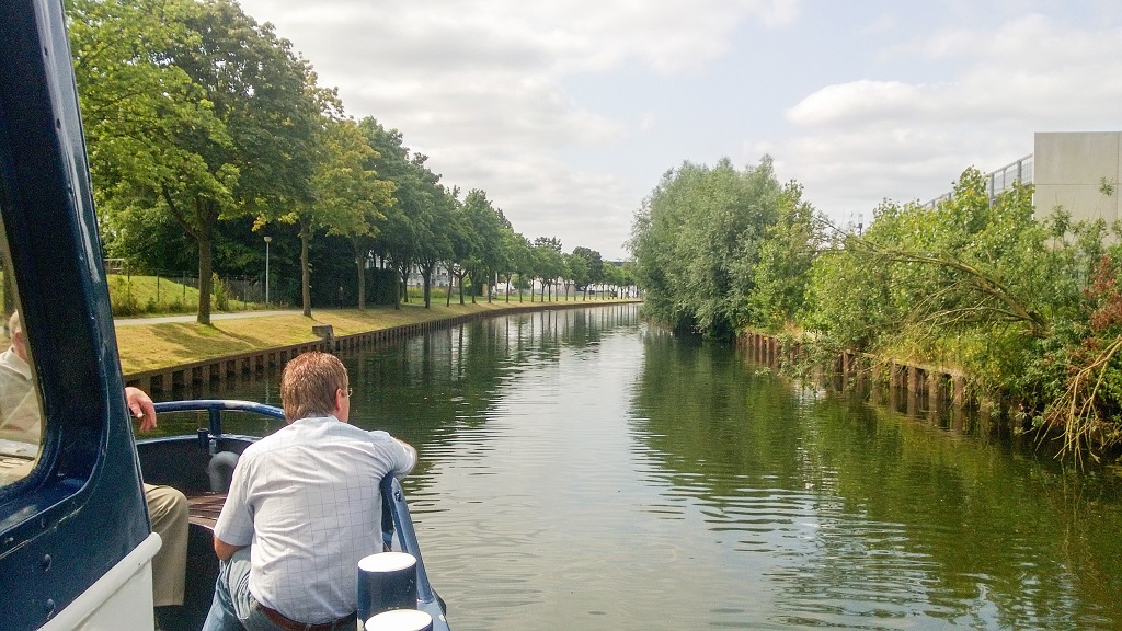 Twee personen zitten achterop een varend schip terwijl het door een smalle, groene waterweg vaart, omgeven door bomen en struiken langs de oevers.