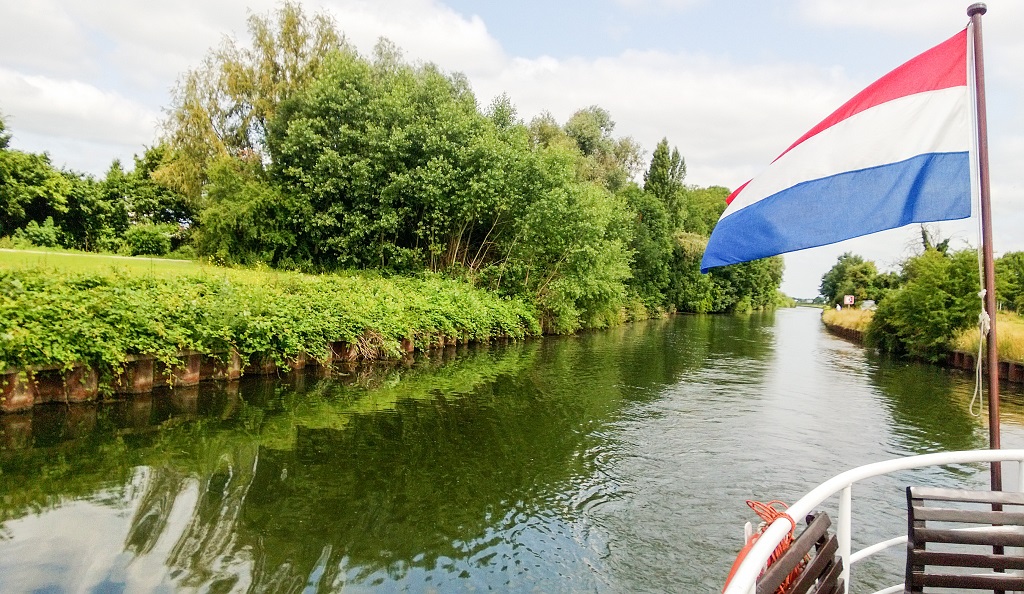 Uitzicht vanaf een varend schip op een smalle groene waterweg, met dichte begroeiing langs de oevers en een wapperende Nederlandse vlag op de voorgrond.
