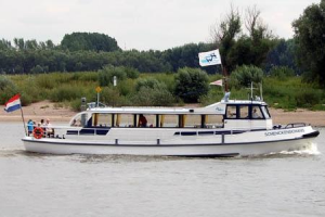 Klein wit passagiersschip met Nederlandse vlag dat over een rivier vaart, met bomen en groen op de achtergrond.