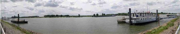 Panoramisch beeld van een rivier met een aanlegsteiger en een groot passagiersschip rechts, omringd door groene oevers onder een bewolkte lucht.