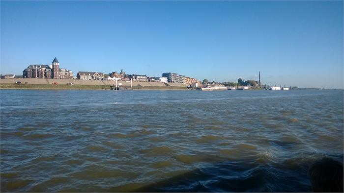 Uitzicht over de Rijn vanaf een schip, met golvend water op de voorgrond en de skyline van een stad met woongebouwen en kade op de achtergrond onder een heldere blauwe lucht