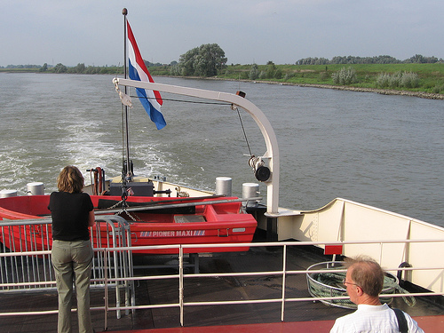 Twee mensen staan op het dek van een rondvaartboot en kijken uit over de rivier met een rood reddingsbootje en een Nederlandse vlag op de achtergrond