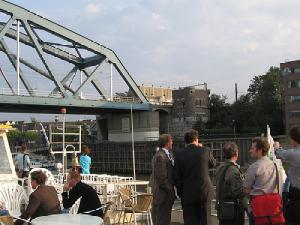 Groep mensen staat op het dek van een rondvaartboot terwijl de boot onder een stalen brug door vaart op de achtergrond zijn gebouwen en een hekwerk zichtbaar