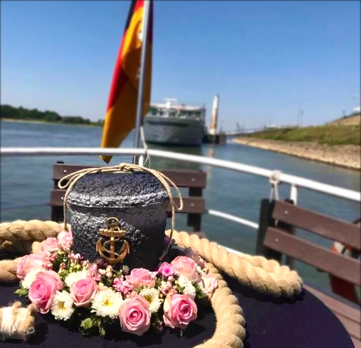 Urn met goudkleurig anker omringd door roze en witte bloemen op een tafel aan boord van een rondvaartschip met zicht op de Rijn en een passerend schip op de achtergrond